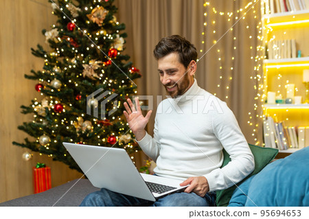 Man at home recording video greetings, Merry Christmas and Happy New Year, sitting on sofa at home near Christmas tree, using laptop for online remote communication with friends, video call. 95694653