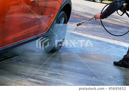 Washing with clean water at self-service car wash. Man washes his orange car at car wash in outdoors. Washing with clean water at self-service car wash. Man washes his orange car at car wash in outdoors. 95695652