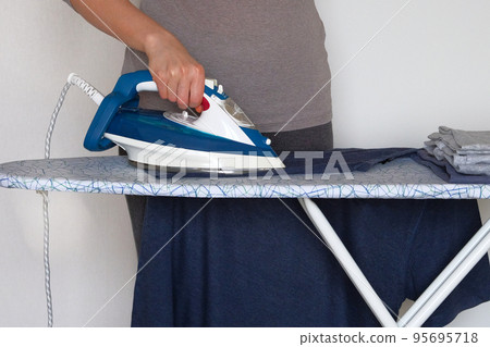 Clothing ironing at home. Young girl is ironing garments at home. Female hand is ironing clothes, close up. Clothing ironing at home. Young girl is ironing garments at home. Female hand is ironing clothes, close up. 95695718