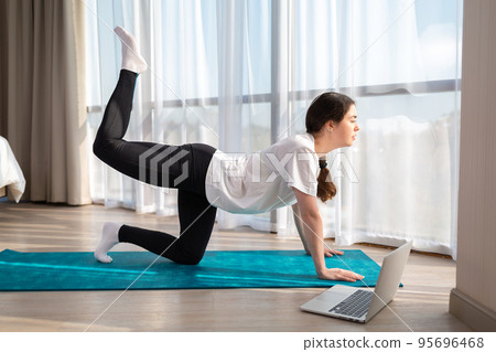 Young beautiful woman in sports clothes, doing yoga online lessons on a laptop. Indoor. Side view. The concept of online training at home Young beautiful woman in sports clothes, doing yoga online lessons on a laptop. Indoor. Side view. The concept of online training at home 95696468