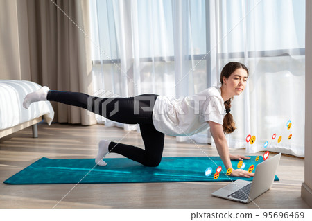 Young woman in sportswear doing yoga live online. Home. Emoticons and emojis fly around the laptop. The concept of social networks, online training and blogging Young woman in sportswear doing yoga live online. Home. Emoticons and emojis fly around the laptop. The concept of social networks, online training and blogging 95696469