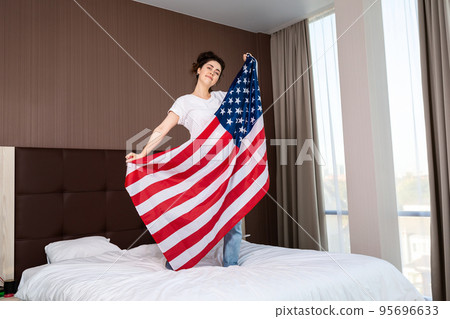 A young smiling woman shows an American flag standing on a bed. Indoor. The concept of the American national holidays and patriotism 95696633
