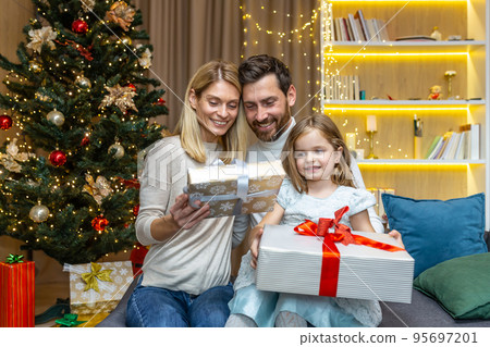 New Year and Christmas holidays, happy family with daughter exchanging gifts and greeting each other sitting on sofa near Christmas tree at home. New Year and Christmas holidays, happy family with daughter exchanging gifts and greeting each other sitting on sofa near Christmas tree at home. 95697201