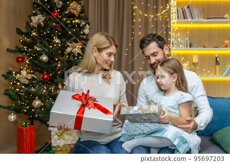 New Year and Christmas holidays, happy family with daughter exchanging gifts and greeting each other sitting on sofa near Christmas tree at home. New Year and Christmas holidays, happy family with daughter exchanging gifts and greeting each other sitting on sofa near Christmas tree at home. 95697203