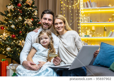 Portrait happy family at home for festivity and new year smiling and looking at camera three people, two adults and a child, using laptop for online shopping in online store, man holding credit card. Portrait happy family at home for festivity and new year smiling and looking at camera three people, two adults and a child, using laptop for online shopping in online store, man holding credit card. 95697902