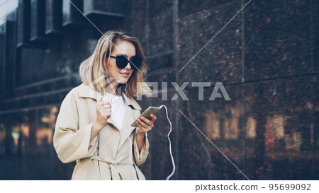 Pretty girl is listening to music through earphones using smartphone and dancing walking in the street and enjoying rhythm and modern city. Girl is wearing sunglasses and trendy coat. 95699092