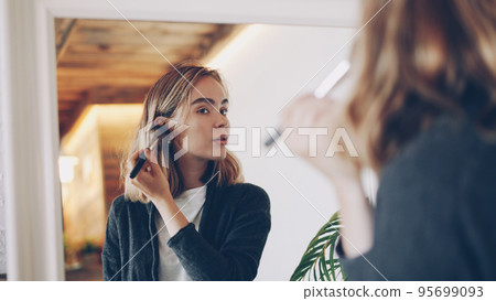 Pretty young girl is finishing make-up touching face and neck with brush then looking in the large mirror and smiling enjoying beautiful look. Focus on mirror reflection. Pretty young girl is finishing make-up touching face and neck with brush then looking in the large mirror and smiling enjoying beautiful look. Focus on mirror reflection. 95699093