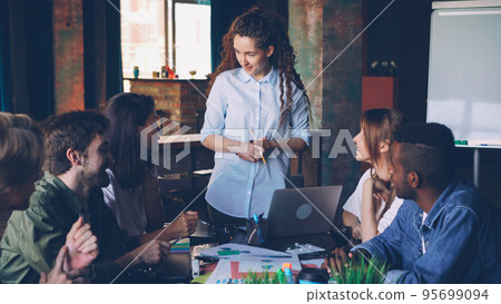 Young woman director of small company is talking to her employees holding business meeting at table in modern loft office. People, teamwork and management concept. Young woman director of small company is talking to her employees holding business meeting at table in modern loft office. People, teamwork and management concept. 95699094