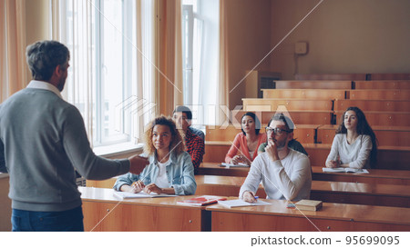 Multi-ethnic group of students is answering teacher's questions, smart young people are talking and smiling sitting in tables while teacher is asking them checking knowledge. Multi-ethnic group of students is answering teacher's questions, smart young people are talking and smiling sitting in tables while teacher is asking them checking knowledge. 95699095