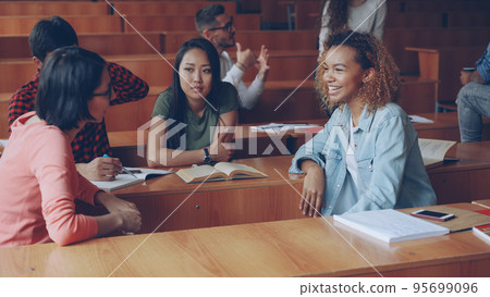 Young women students are having conversation after lectures at college, girls are talking and gesturing while other students are chatting in background. Young and communication concept. Young women students are having conversation after lectures at college, girls are talking and gesturing while other students are chatting in background. Young and communication concept. 95699096