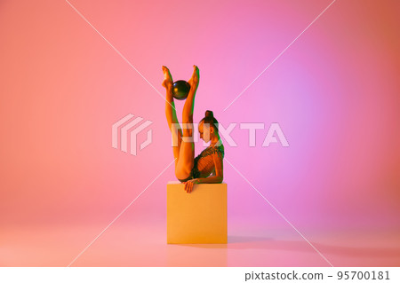 Studio shot of young charming girl, rhythmic gymnast training with sports equipment isolated over pink background in neon light filter 95700181
