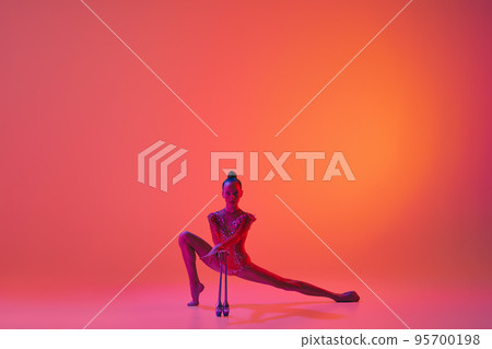 Studio shot of young charming girl, rhythmic gymnast training with sports equipment isolated over pink background in neon light filter Studio shot of young charming girl, rhythmic gymnast training with sports equipment isolated over pink background in neon light filter 95700198