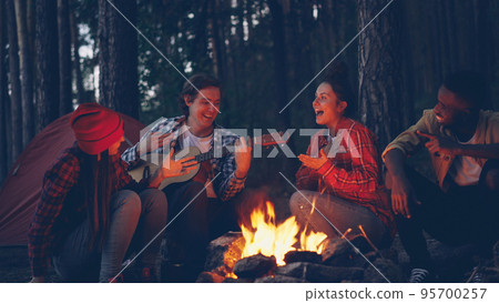 young man tourist is playing the guitar while his friends are singing and laughing sitting around fire in the wood in the evening enjoying nature and good company. young man tourist is playing the guitar while his friends are singing and laughing sitting around fire in the wood in the evening enjoying nature and good company. 95700257