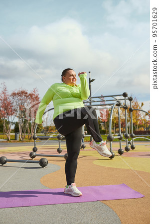 Happy overweight young woman doing fitness exercises at street public sports ground, outdoors. Sunny autumn day. Sport, health, mood, body positive Happy overweight young woman doing fitness exercises at street public sports ground, outdoors. Sunny autumn day. Sport, health, mood, body positive 95701629