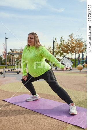 Happy overweight young woman doing fitness exercises at street public sports ground, outdoors. Sunny autumn day. Sport, health, mood, body positive Happy overweight young woman doing fitness exercises at street public sports ground, outdoors. Sunny autumn day. Sport, health, mood, body positive 95701705
