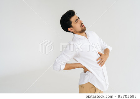 Studio portrait of unhealthy young man in shirt touching injured sore back, suffering lower lumbar discomfort, muscle pain of overwork, pinched nerve and grimacing on white isolated background. 95703695