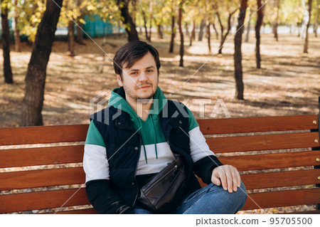 Looking at camera young caucasian unshaven man 34 years old man in sweatshirt and warm vest sits on bench in autumn park. Outdoor park, sunny day, waist up lifestyle portrait. Waiting concept 95705500