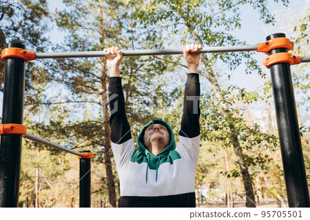 Young caucasian unshaven man 34 years old doing pull ups at the gym, unsporting non- athletic man hanging on bars on sunny autumn day. Outdoor park sports equipment. Lifestyle portrait. 95705501