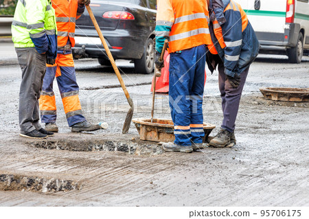 Road workers in orange overalls are repairing old sewer manholes on the roadway against the backdrop of passing cars. Copy space. Road workers in orange overalls are repairing old sewer manholes on the roadway against the backdrop of passing cars. Copy space. 95706175