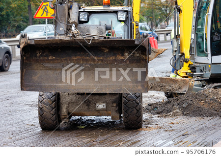 A wide metal bucket of a road bulldozer is raised above the surface of the road section being repaired. A wide metal bucket of a road bulldozer is raised above the surface of the road section being repaired. 95706176