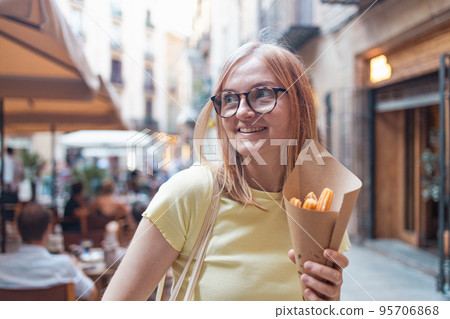 Close-up of young blonde woman eating eating traditional spanish snack named Churros with sugar and chocolate sauce, a fried-dough pastry based snack. Close-up of young blonde woman eating eating traditional spanish snack named Churros with sugar and chocolate sauce, a fried-dough pastry based snack. 95706868