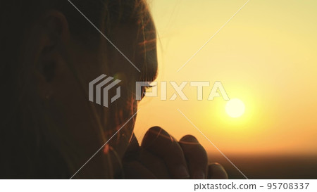 girl prays looking sunset, long hair flying away in glare sunlight rays strong wind, looking at dawn, lonely hike of brave girl, looking into sky with her eyes, believing good. close-up hands face. 95708337