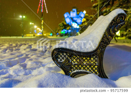 Winter park in the evening covered with snow with a row of lamps View of bench and shining lantern through snowing. Winter night landscape - evening in the night snowy park with lonely benches 95717599