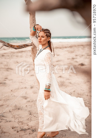 Model in boho style in a white long dress and silver jewelry on the beach. Her hair is braided, and there are many bracelets on her arms. Model in boho style in a white long dress and silver jewelry on the beach. Her hair is braided, and there are many bracelets on her arms. 95718970