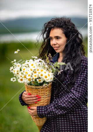 A woman stands on a green field and holds a basket with a large bouquet of daisies in her hands. In the background are mountains and a lake. A woman stands on a green field and holds a basket with a large bouquet of daisies in her hands. In the background are mountains and a lake. 95718974