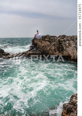 A woman in a storm sits on a stone in the sea. Dressed in a white long dress, waves crash against the rocks and white spray rises. A woman in a storm sits on a stone in the sea. Dressed in a white long dress, waves crash against the rocks and white spray rises. 95718975