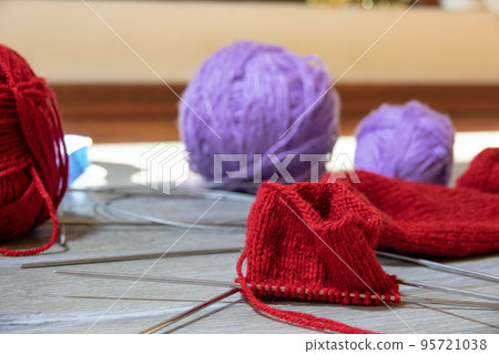 Sock in the process of knitting with needles and yarn of red color on a gray wooden table. Clews threads of red and lilac color on a blurred background 95721038