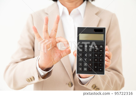 A middle-aged woman in a suit holding a calculator and making a circle sign A middle-aged woman in a suit holding a calculator and making a circle sign 95722316