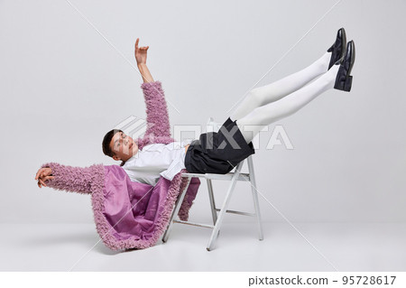 Portrait of stylish boy in pink furry coat, white tights, black shorts lying on chair, posing isolated over grey background. Freedom Portrait of stylish boy in pink furry coat, white tights, black shorts lying on chair, posing isolated over grey background. Freedom 95728617