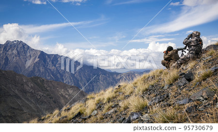 Couple of people dressed in camouflage are carefully examining the mountainous terrain with binoculars. Couple of people dressed in camouflage are carefully examining the mountainous terrain with binoculars. 95730679