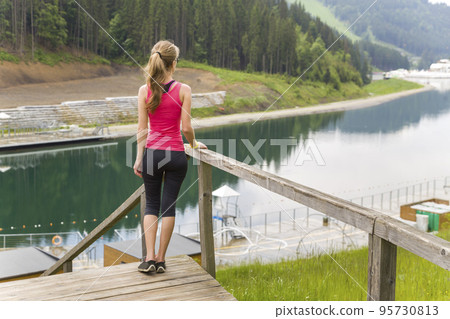Back view of a young sportive woman in fitness clothes standing outdoors near big lake in mountains enjoying nature view. 95730813