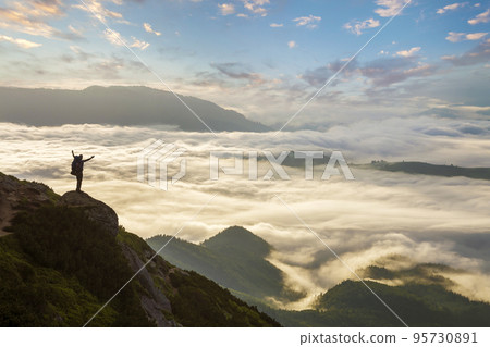 Wide mountain panorama. Small silhouette of tourist with backpack on rocky mountain slope with raised hands over valley covered with white puffy clouds. Beauty of nature, tourism and traveling concept 95730891