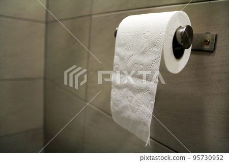 Close-up of white soft tissue paper roll in bathroom, restroom, wc on light brown beige tiling background ion simple shiny chrome holder. Modern lifestyle, hygiene, conviencies. 95730952