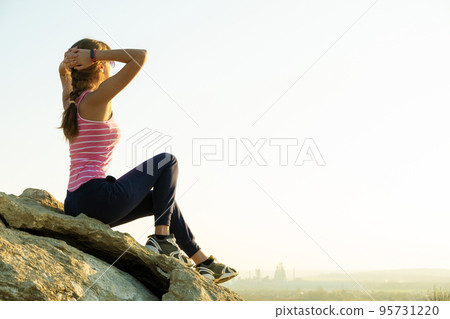 Woman hiker sitting on a steep big rock enjoying warm summer day. Young female climber resting during sports activity in nature. Active recreation in nature concept. 95731220