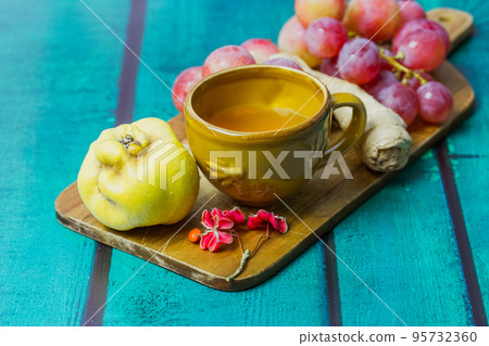 Beautiful fall still life with yellow cup on saucer. Bunch of red grapes, ripe agave, ginger root lie on cutting board. Fallen oak, maple leaves. Wooden green emerald background with space for text. 95732360