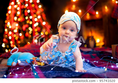 Small baby girl in pajama playing with garland while crawling on checkered plaid near Christmas tree 95741020