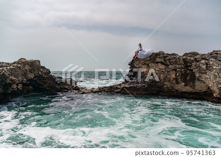 A woman in a storm sits on a stone in the sea. Dressed in a white long dress, waves crash against the rocks and white spray rises. 95741363