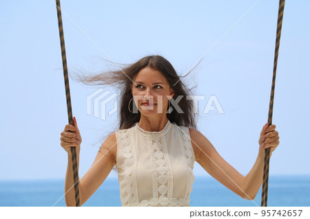Beautiful young woman rides a swing in front of the ocean in Thailand. The happy girl is smiling, looking away and enjoying her vacation. Beautiful young woman rides a swing in front of the ocean in Thailand. The happy girl is smiling, looking away and enjoying her vacation. 95742657