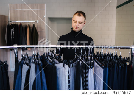 Handsome customer in black golf try to fing some clother for new season. Autumn and winter clothes.. Front view throught shelves Handsome customer in black golf try to fing some clother for new season. Autumn and winter clothes.. Front view throught shelves 95745637