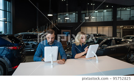 Caucasian married couple chooses a car in a car dealership on digital tablets.  95752825