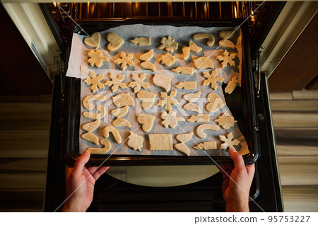 Top view: hands of confectioner putting a baking sheet with carved molds of gingerbread dough in the hot oven. Housewife preparing delicious homemade biscuits for Christmas, according to family recipe 95753227