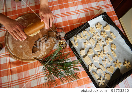 Top view. Selective focus. Housewife's hands roll out dough with a rolling pin, a baking sheet with carved molds of gingerbread dough on the table. Preparing homemade Christmas pastries in the kitchen Top view. Selective focus. Housewife's hands roll out dough with a rolling pin, a baking sheet with carved molds of gingerbread dough on the table. Preparing homemade Christmas pastries in the kitchen 95753230