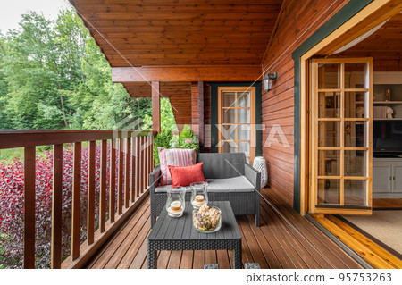 Cozy terrace with table and sofa at wooden cottage house surrounded by greenery and trees in countryside on cloudy summer day Cozy terrace with table and sofa at wooden cottage house surrounded by greenery and trees in countryside on cloudy summer day 95753263
