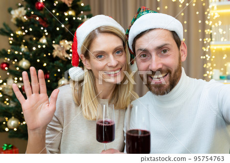 Family in love couple man and woman together for Christmas greeting friends talking remotely on video call looking at smartphone camera in Santa Claus hats with glasses of wine, online greetings. Family in love couple man and woman together for Christmas greeting friends talking remotely on video call looking at smartphone camera in Santa Claus hats with glasses of wine, online greetings. 95754763