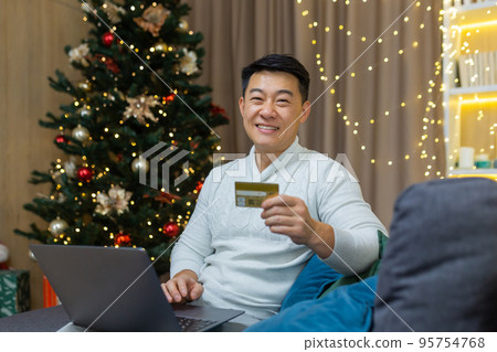 Man with bank credit card smiling and looking at camera with laptop sitting on sofa in living room at home near Christmas tree, portrait of happy asian shopper in online store for new year. Man with bank credit card smiling and looking at camera with laptop sitting on sofa in living room at home near Christmas tree, portrait of happy asian shopper in online store for new year. 95754768