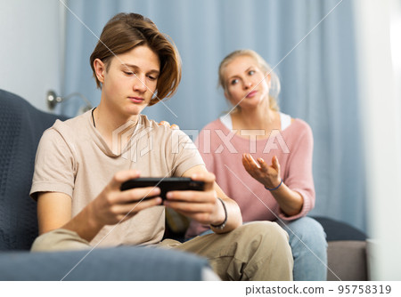 Teenager playing game on phone while upset mother talking to him Teenager playing game on phone while upset mother talking to him 95758319
